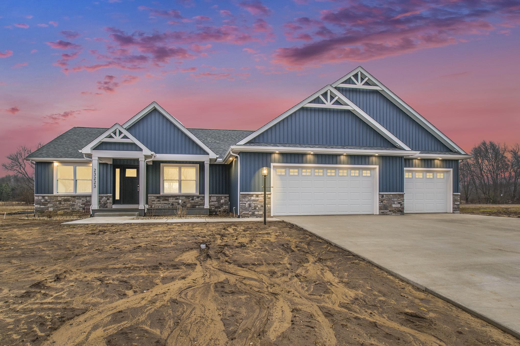 Blue farmhouse with two garages with muddy lawn for new grass against pink and purple sky.